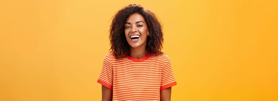 Waist-up shot of outgoing happy charming dark-skinned female with curly hairstyle laughing joyfully posing in striped trendy t-shirt over orange background enjoying nice casual conversation. Lifestyle.