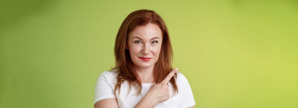 Kind cheerful good-looking middle-aged woman 50s redhead. white t-shirt smiling modest assured give advice pointing upper left corner indicating great promotion advertising product green background.