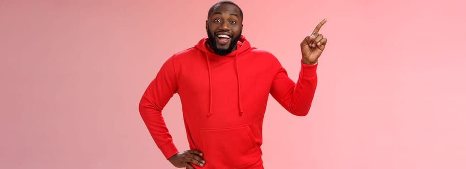 Enthusiastic happy good-looking young black guy beard pointing upper left corner smiling approval like curious about interesting item sale ask question excited impressed, standing pink background.