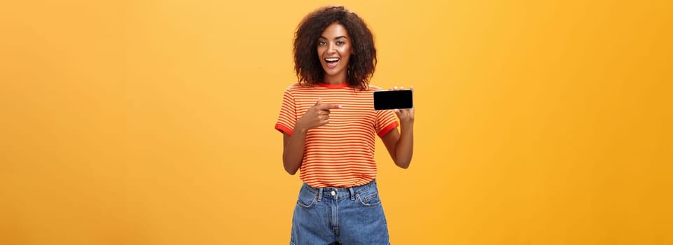 Girl showing picture in phone bragging about her cool vacation. Charming friendly and sociable african american woman with curly hair holding cellphone pointing at smartphone screen over orange wall. Technology and advertisement concept