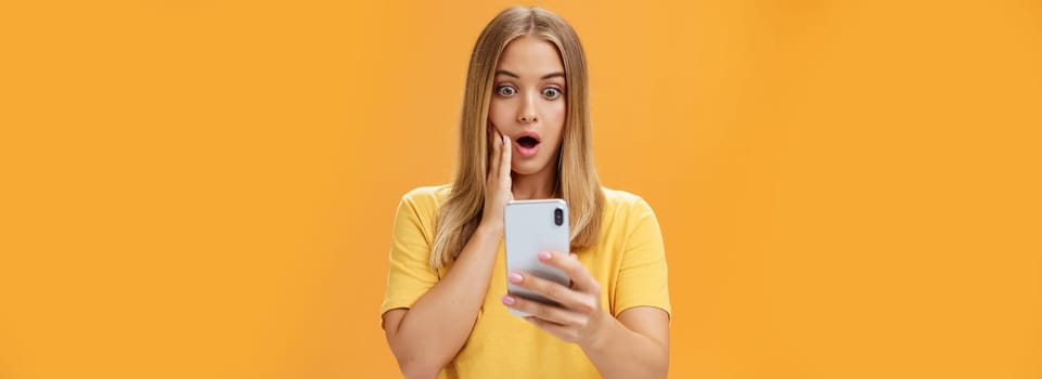 Waist-up shot of shocked and stunned emotive young woman reacting to shocking terrible news reading online from internet gasping opening mouth nervously touchin cheek staring at smartphone screen. Technology concept