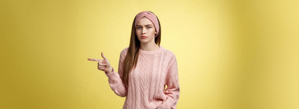 Pissed puzzled young attractive arrogant moody girlfriend in sweater, headband looking irritated, intense frowning pointing finger at copy space, directing person sits on her place, annoyed.
