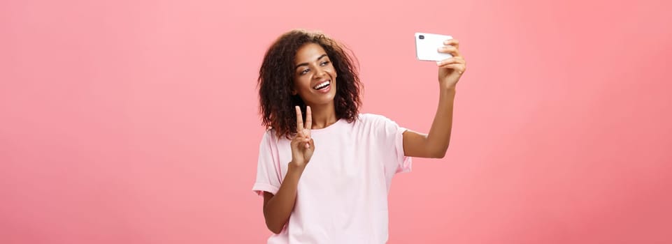 Stylish sociable good-looking dark-skinned female student with curly hairstyle pulling hand with smartphone near face taking selfie showing peace sign to device screen while smiling carefree. Lifestyle.