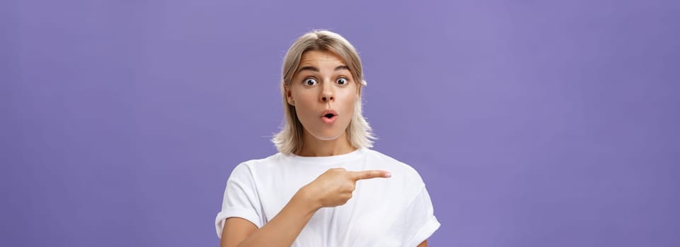 Waist-up shot of overwhelmed excited and surprised attractive urban female in white t-shirt folding lips gasping from amazement being questioned asking question and pointing left over purple wall. Copy space