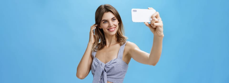 Attractive woman with good self-esteem in stylish matching outfit flicking hair behind ear smiling joyfully at smartphone screen, taking selfie to post in internet and gain followers over blue wall. Lifestyle concept