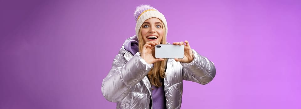 Amused fascinated female in silver jacket head smiling astonished excited look forward holding smartphone recording video taking shots famous person mobile phone camera, purple background.