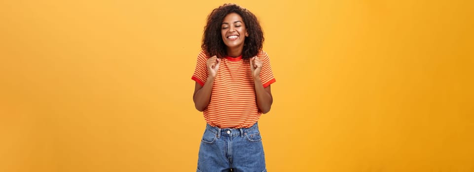 Girl eager to see lover coming from trip standing excited and thrilled with happy broad smile clenching fists grinning with closed eyes feeling joyful and amused over orange background. Lifestyle.