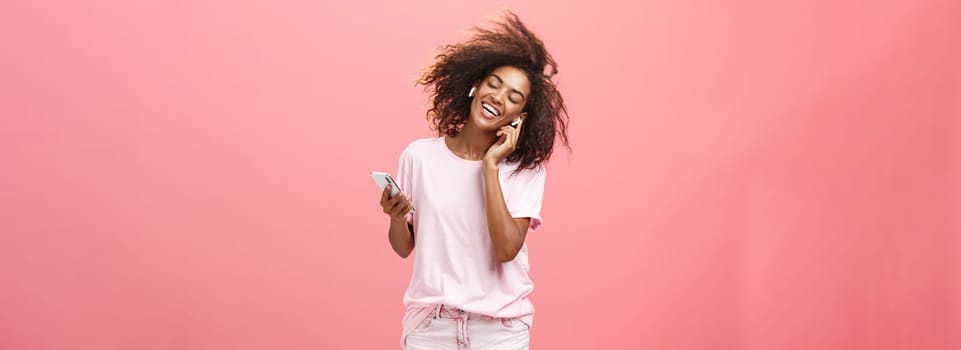 Getting rid of worried with good tunes in ears. Pleased attractive young african woman in trendy outfit listening music in wireless earphones touching earbud smiling dancing and holding smartphone. Technology concept