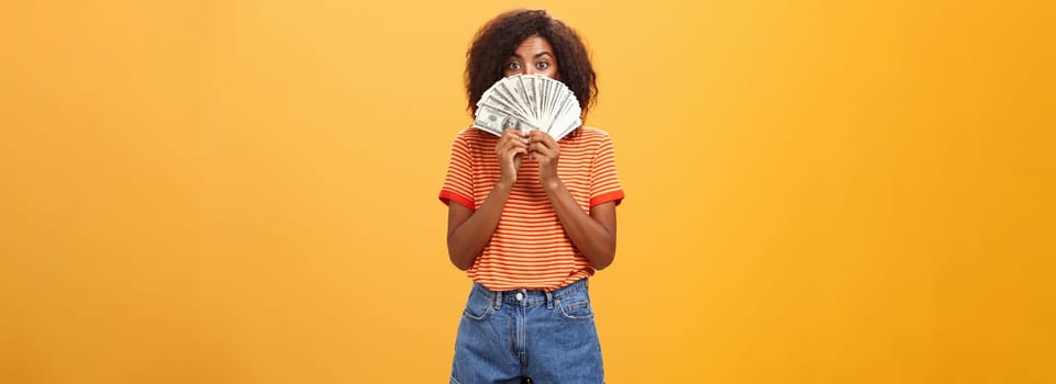 Woman gonna waste lots of cash today. Pleased and delighted rich african american girl with curly hairstyle hiding face behind bunch of money peeking mysteriously at camera winning millions. Copy space
