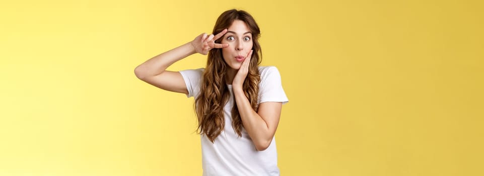 Silly coquesttish good-looking caucasian girl curly hairstyle folding lips amused interested touch cheek feminine sensual pose show victory peace sign upbeat stand intrigued yellow background. Lifestyle.