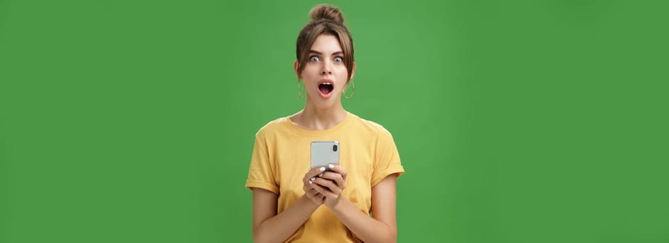 Portrait of shocked speechless and impressed beautiful white girl with combed hair in yellow t-shirt holding smartphone, dropping jaw from excitement reacting to cool app over green background.