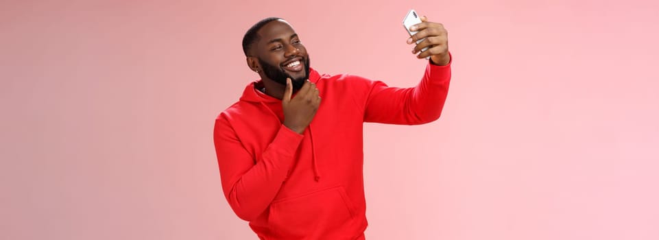 Lifestyle. Cheeky cool african american confident bearded guy in red hoodie taking selfie update date app profile photo holding smartphone raised touching face sassy smiling look phone display flirty.