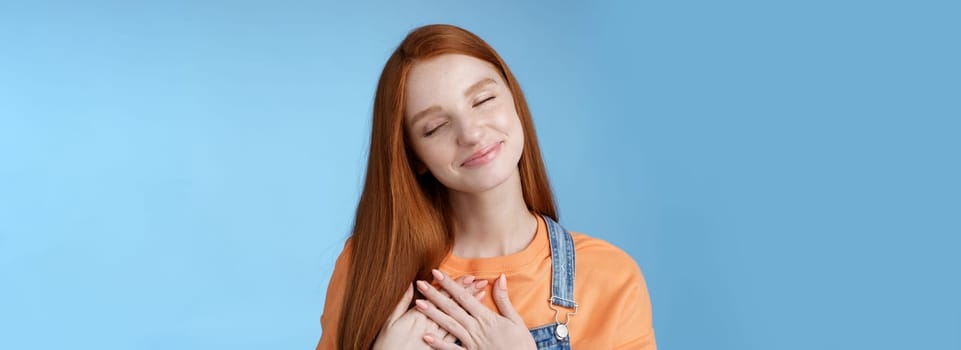 Dreamy touched romantic ginger girlfriend close eyes recalling heartwarming romance touch heart palms pressed chest smiling tenderly feel love care sympathy express affection, blue background.
