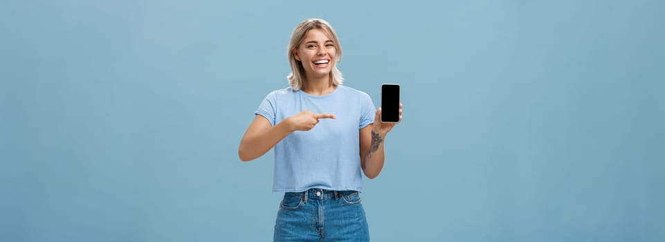 Look at this hilarious photo. Entertained attractive happy woman with fair hair in casual t-shirt and denim shorts showing smartphone at camera pointing at device screen smiling broadly over blue wall.