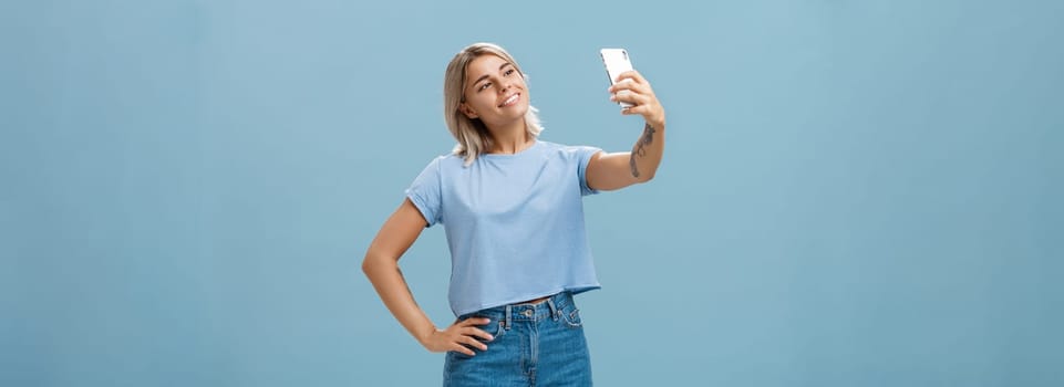 Lifestyle. Stylish cute female gym manager taking selfie with new smartphone posing with broad pleased smile and holding hand on hip making post for followers standing carefree and relaxed over blue wall.