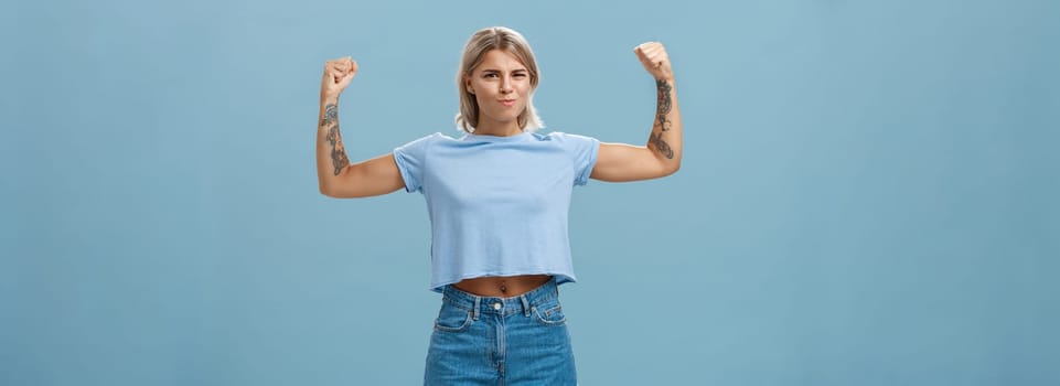 Lifestyle. Strong and powerful good-looking sportswomen with tattoos in t-shirt and shorts raising arms showing big muscles and biceps smiling proudly while bragging with physical strength over blue wall.