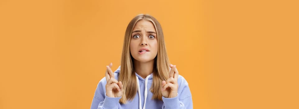 Worried anxious hopeful young woman with fair hair in stylish over-sized hoodie biting lower lip frowning concerned crossing fingers for good luck praying for dream come true against orange wall. Lifestyle.