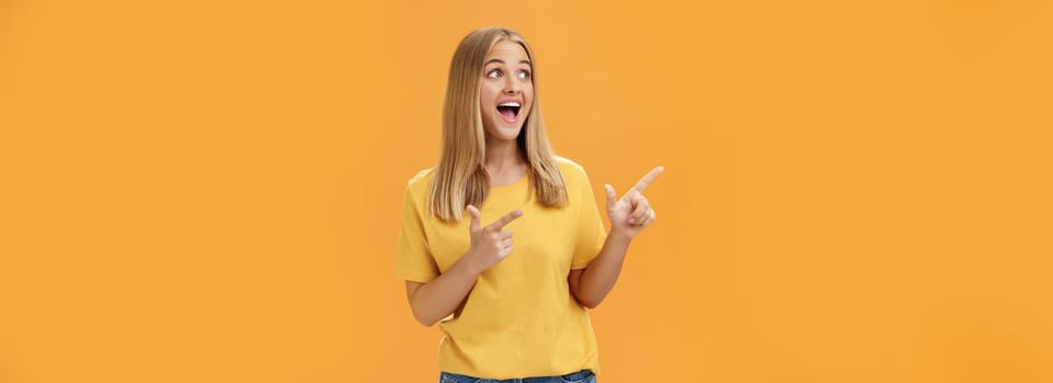 Amused enthusiastic and charismatic girl with blond hair and tanned pure skin without makeup pointing and looking at upper left corner with joyful happy smile over orange background. Body language concept