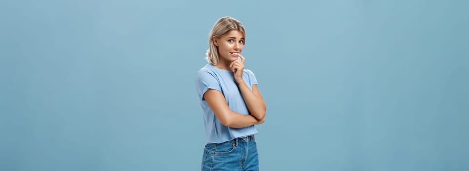 Studio shot of unconfident silly and insecure cute feminine girl with blond hair standing half-turned over blue background holding finger on lip while being shy ask question smiling hesitating. Emotions concept