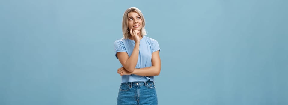 Dreamy thoughtful and creative artistic blonde woman in denim shorts and t-shirt smiling curiously touching lip and gazing at upper left corner feeling nostalgic, thinking over blue wall.