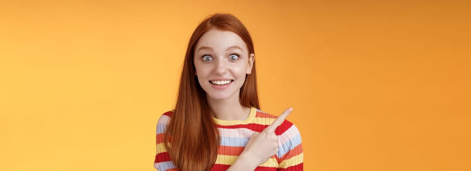 Excited thrilled good-looking young silly redhead girl surprised pop eyes camera gasping impressed pointing upper left corner see super star grinning excitement delight, orange background. Copy space