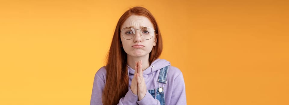 Please beg you. Clingy upset sad caucasian redhead girlfriend wearing glasses frowning grimacing pouting pleading help asking favour need urgently borrow something, orange background.