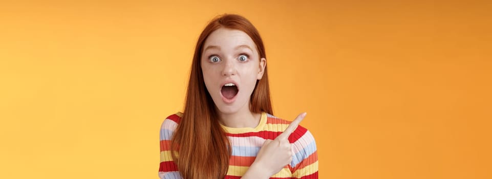 Impressed speechless young redhead 20s girl drop jaw gasping amused wide eyes staring camera surprised see awesome stunning new product pointing left index finger questioned, orange background.