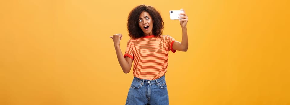 Woman recording video blog pointing at strange object behind her. Portrait of concerned and curious stylish famous internet star holding smartphone talking in cellphone camera over orange background. Technology concept