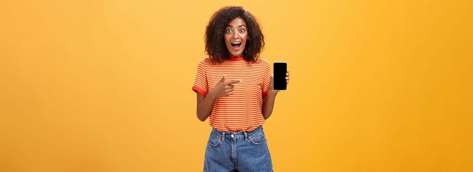 Impressed fascinated stylish slim african american curly-haired woman in striped t-shirt dropping jaw from amazement holding awesome smartphone pointing at device screen over orange wall. Lifestyle.