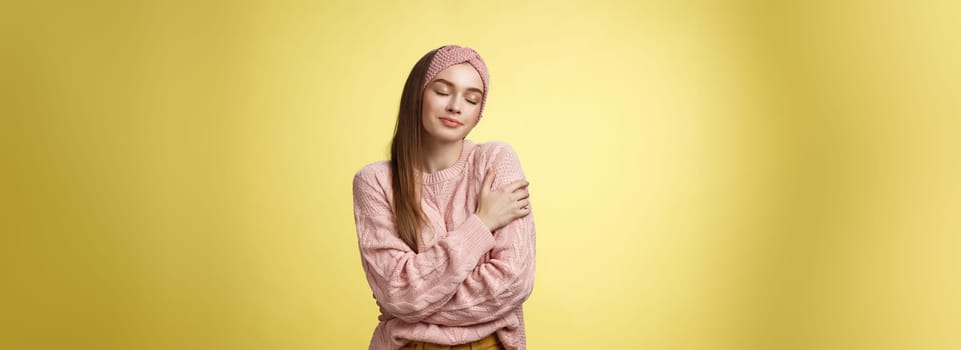 Sweet glamour young girl wearing warm comfy sweater embracing herself crossing arms over body in hug, smiling soft and kind close eyes, recalling lovely days, tender memories over yellow wall.