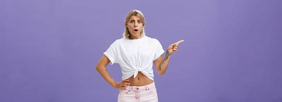Girl being unhappy with unfair thing happened. Shocked displeased complaining, confused woman with tanned skin blond hair and tattoos opening mouth and frowning from jealousy or regret pointing left.