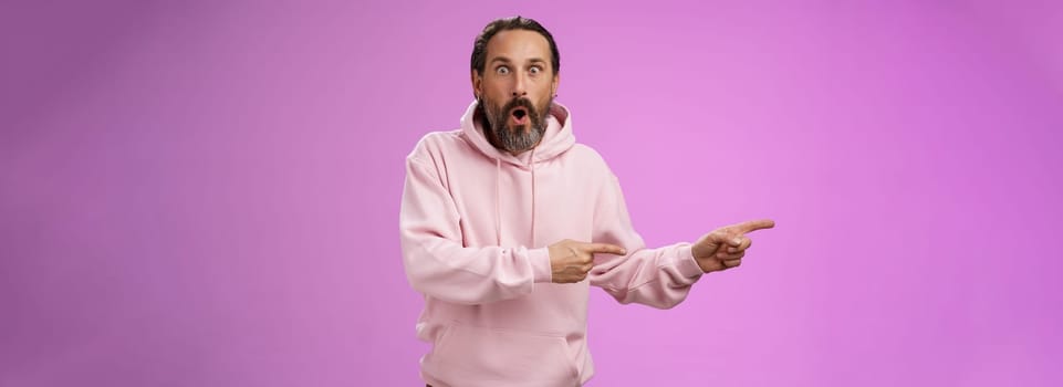Astonished fascinated curious mature bearded man with grey hair in pink hoodie folding lips amazed pointing left index fingers widen eyes camera impressed thrilled wanna go check out cool promo. Lifestyle.