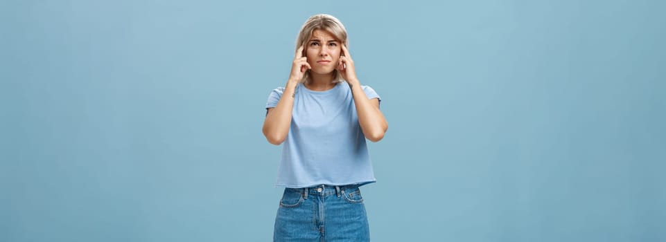 Lifestyle. Girl cannot concentrate hearing loud noise comming from upstairs frowning feeling intense and dissatisfied closing ears with index fingers looking up while complaining on awful sound over blue wall.