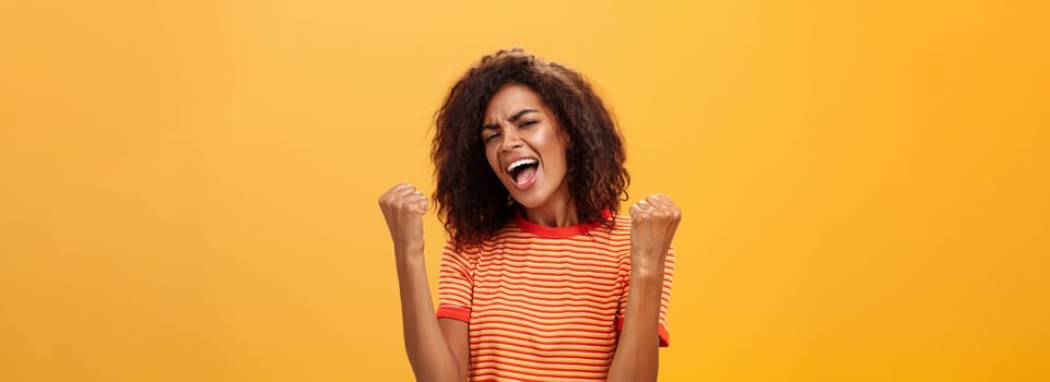 Cheerful delighted and enthusiastic african american woman with afro hairstyle clenching raised fists yelling yes from triumph and joy of success standing satisfied of victory over orange wall. Lifestyle.