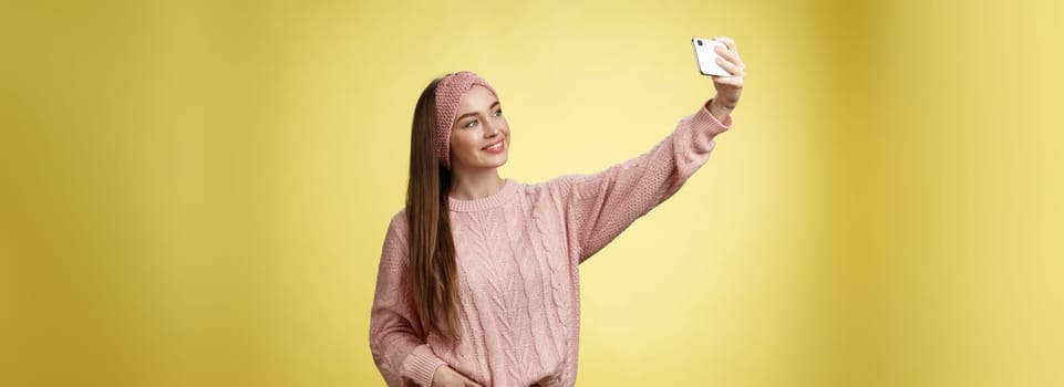 Popular glamour young female internet lifestyle blogger taking selfie on new smartphone extending arm taking picture herself against yellow background smiling at cellphone screen, posing cheeky.
