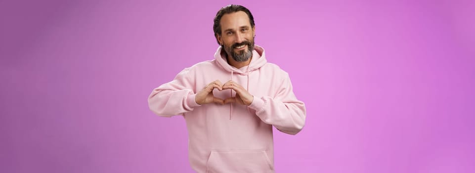 Family in heart. Portrait lovely handsome romantic bearded man in pink hoodie passionatly looking camera show love gesture smiling cute expressing romantic sympathy attitidue, purple background.