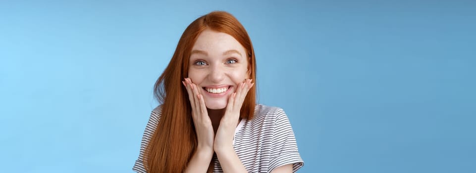 Excited happy cheerful redhead ginger girl smiling joyfully touch cheeks surprised receive amazing perfect b-day gift standing astonished triumphing rejoicing standing blue background. Copy space