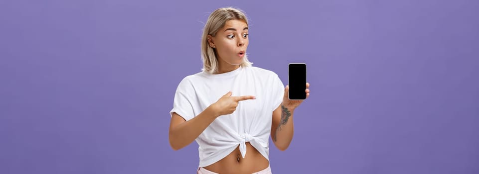 Waist-up shot of impressed and delighted gorgeous european female with blond hair and tattoos looking and pointing at awesome brand new smartphone standing over purple background. Technology concept