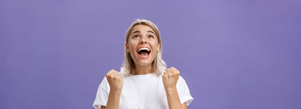 Waist-up shot of grateful delighted lucky girl with attractive tan in white t-shirt clenching fist from positive emotions gazing delighted and happy up thankful giving thanks god for fulfilling dream. Emotions concept