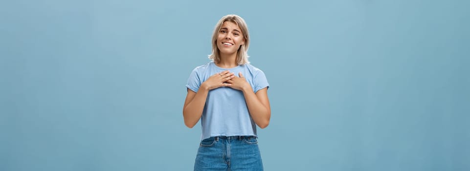 So sweet thanks. Touched beautiful and emotive blond girl in t-shirt and shorts with tanned skin holding palms on breast, sighing and smiling joyfully being thankful and grateful over blue wall. Emotions concept