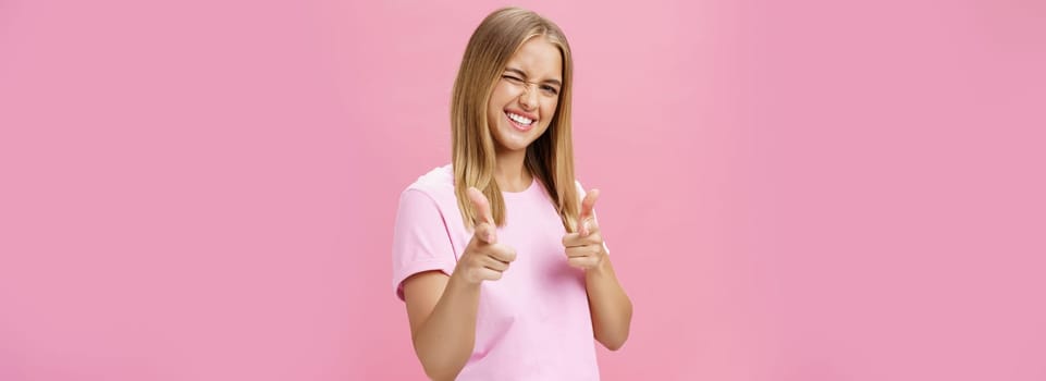 Woman expressing positive attitude towards camera pointing with fingers and winking joyfully smiling being uplifted, standing in good mood with optimistic gestures against pink background. Copy space