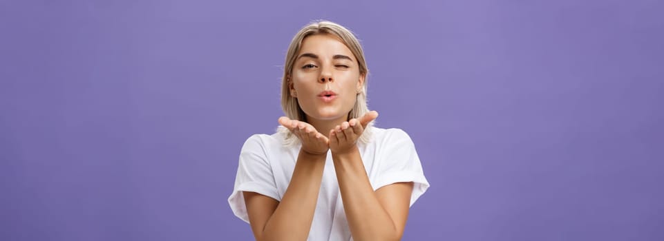 Blowing sweet kiss to all fans. Flirty tender and cute stylish european female with blond hair and tanned skin holding palms near mouth folding lips while sending mwah at camera over purple wall.