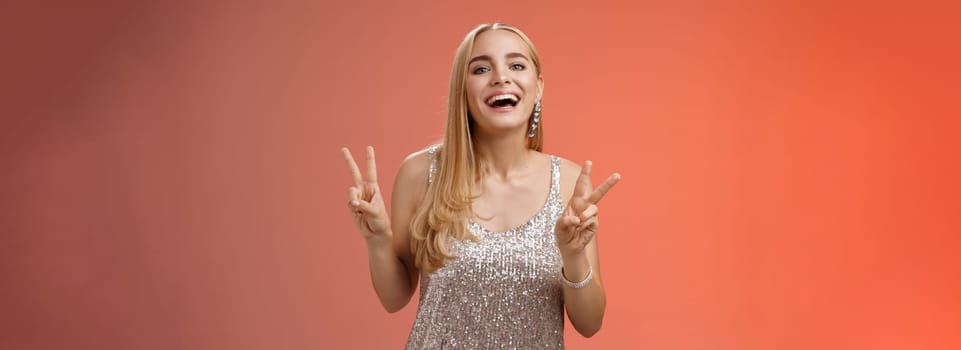 Funny carefree charming caucasian blond female model in silver stylish glamour dress show peace victory gesture smiling broadly have fun celebrating birthday party, standing red background.