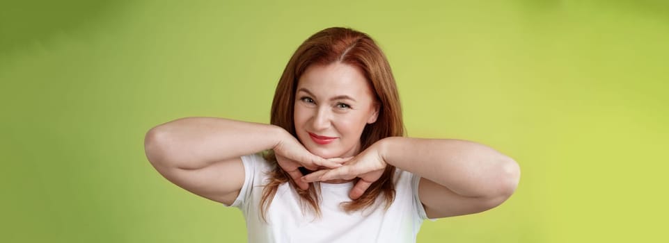 Aging, cosmetology, wellbeing concept. Happy self-assured redhead woman hold hands under jawline smiling showing facial blemished self-accepting wrinkles applying skincare product green background.