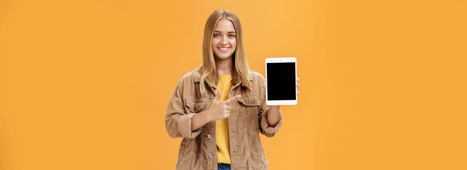 Get ready for autumn semister with new digital tablet. Portrait of charming female in corduroy jacket showing gadget screen pointing at device and smiling pleased and friendly over orange wall. Technology and advertising concept