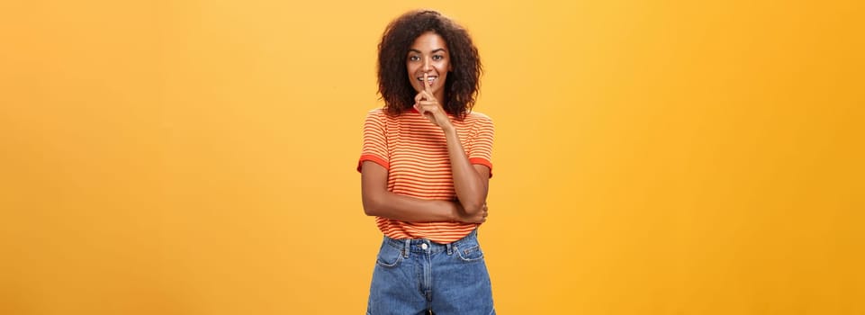 Keep secret inside I trust you. Feminine and sensual good-looking stylish dark-skinned woman with curly hair in trendy outfit shushing at camera with delighted mysterious look hiding something. Body language concept