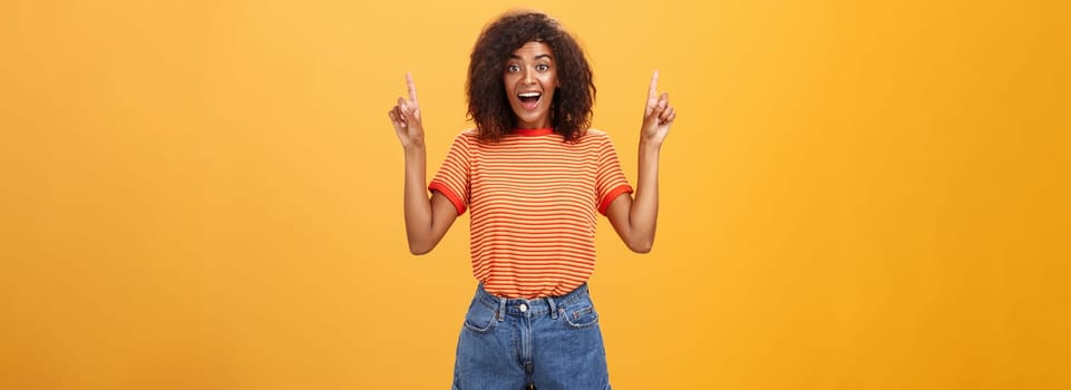 Charmed and impressed african american stylish woman seeing incredible and awesome item on sale raising hands and pointing up with broad grin being excited of new purchase over orange wall. Emotions and advertisement concept