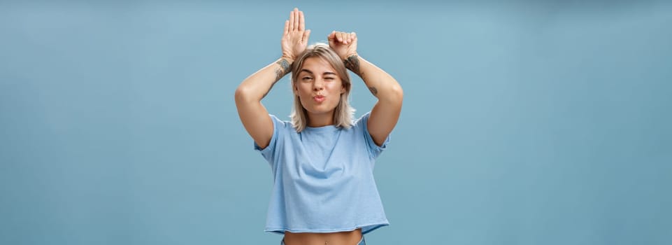 Lifestyle. Flirty attractive and stylish cool girlfriend with tattooed arms and pierced belly winking folding lips in air kiss showing bunny ears with palms on head standing over blue background playful.