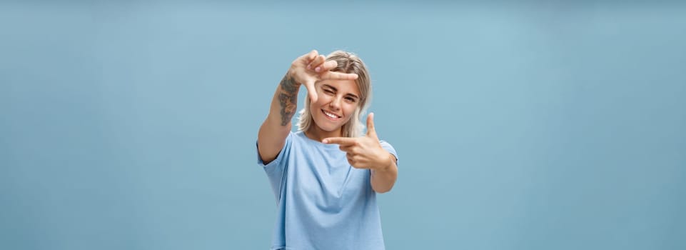 Be creative and happy. Portrait of charming enthusiastic female designer with blond hair and tattoo on arm winking and smiling joyfully making frame gesture as if measuring space over blue background.