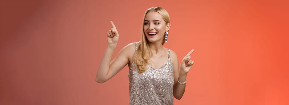 Carefree elegant joyful charming blond girlfriend dancing boyfriend party nightclub standing red background in silver glittering dress raising hands up moving body music rhythm, smiling.